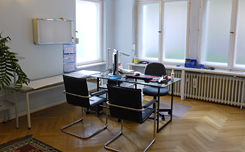 Das Foto zeigt den zweiten der drei Behandlungsräume, mit Parkettboden, Glasschreibtisch und Stahlrohrstühlen.